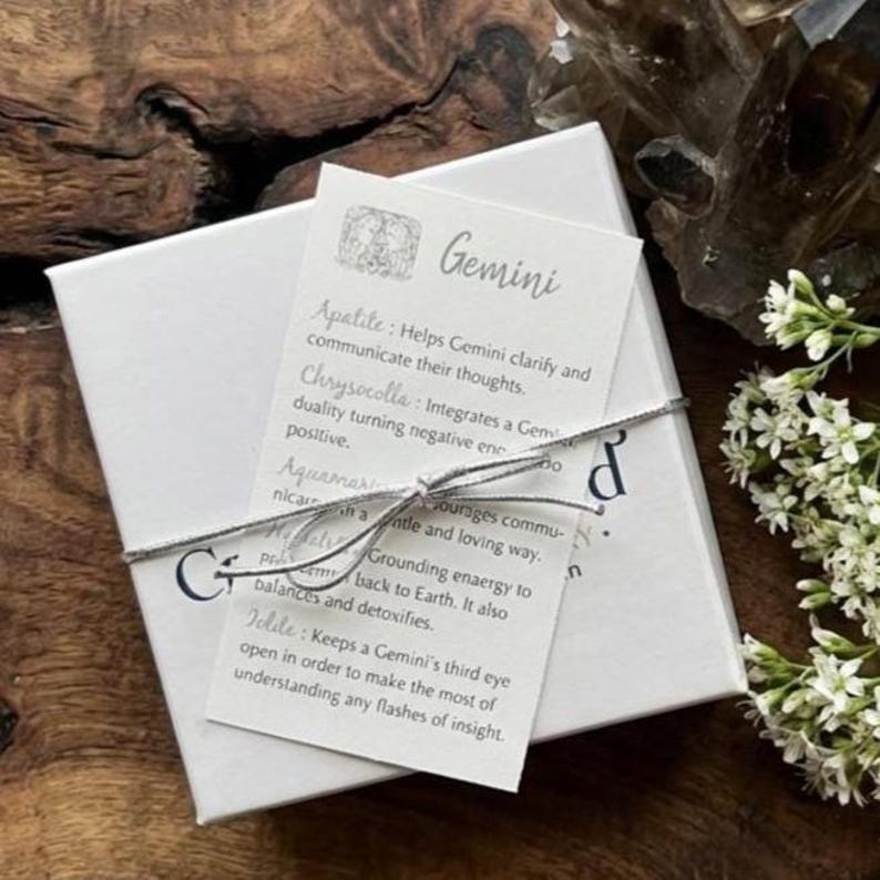 May include: A white box tied with silver string, featuring a Gemini card with text about gemstones. The card lists gemstones like Apatite, Chrysocolla, and Aquamarine. The box is on a wooden surface, with flowers and crystals in the background.