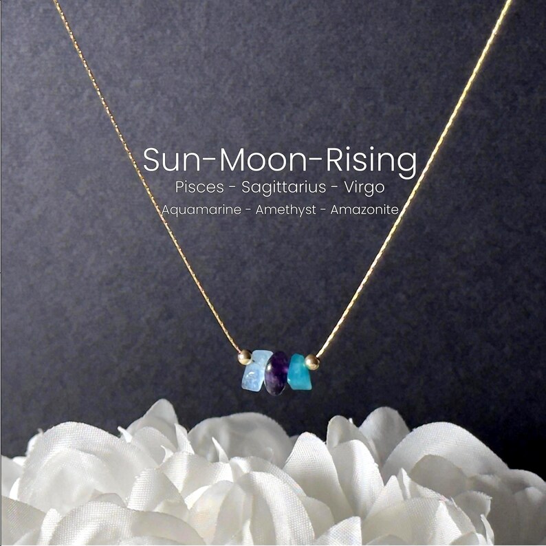 May include: A gold necklace with three gemstones in shades of blue and purple. The necklace is set against a dark backdrop, with the text "Sun-Moon-Rising" and astrological signs. The necklace is displayed over white flowers.