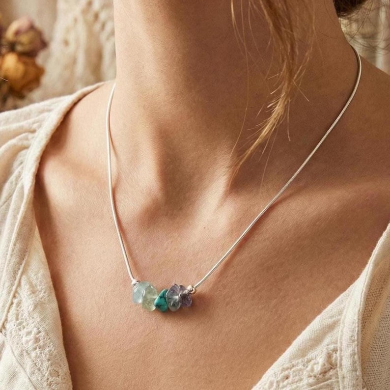 May include: A silver-toned snake chain necklace featuring a central cluster of four small, raw, tumbled gemstone beads in shades of translucent green, teal, and purple, resting against the collarbone on a cream-colored textured fabric.
