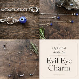 May include: Various necklaces and chains with silver and gold tones, featuring blue evil eye charms and gemstone accents. The image includes the text "Optional Add-On Evil Eye Charm". The jewelry is displayed on a dark wooden surface.