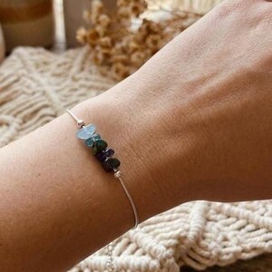 May include: A delicate silver bracelet featuring a row of colorful gemstones. The stones include shades of blue, green, and purple. The bracelet is worn on a wrist, showcasing the jewelry's design and color.