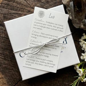 Pu&ograve; includere: Una scatola regalo quadrata bianca legata con un sottile cordoncino argentato, sormontata da un cartoncino rettangolare con un'illustrazione di leone e il testo Leo. Il cartoncino elenca propriet&agrave; dei cristalli come Carnelian, Citrine, Labradorite, Garnet e Peridot.