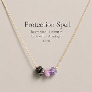 May include: A gold-toned necklace featuring a cluster of five small, irregular tumbled gemstones in shades of black, grey, purple, and light blue. The text Protection Spell, Tourmaline, Hematite, Lepidolite, Amethyst, and Iolite is printed above.