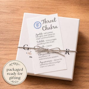 May include: A white gift box tied with silver string, featuring a card labeled "Throat Chakra." The card lists properties of gemstones like Apatite and Aquamarine. A round sticker reads "packaged ready for gifting."