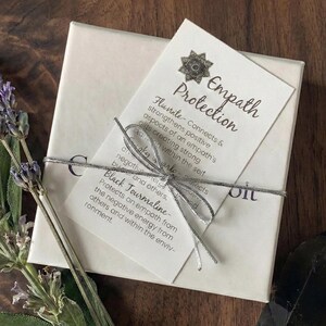 May include: A square white gift box tied with a silver metallic string, featuring a card titled Empath Protection. The card includes text about Fluorite, Clear Quartz, and Black Tourmaline, decorated with a small silver star-shaped charm.