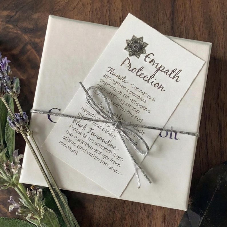 May include: A square white gift box tied with a silver metallic string, featuring a card titled Empath Protection. The card includes text about Fluorite, Clear Quartz, and Black Tourmaline, decorated with a small silver star-shaped charm.