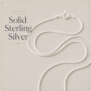 May include: A thin, flexible silver snake chain necklace featuring a circular spring ring clasp. The text on the left side reads: MATERIAL Solid Sterling Silver. The chain is arranged in a curved, looping pattern against a neutral background.