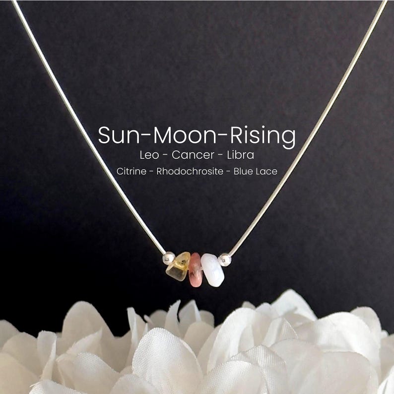 May include: A silver necklace with three gemstones in yellow, pink, and white. The necklace has the text "Sun-Moon-Rising" and astrological signs. The gemstones are citrine, rhodochrosite, and blue lace. The necklace is displayed against a dark background.