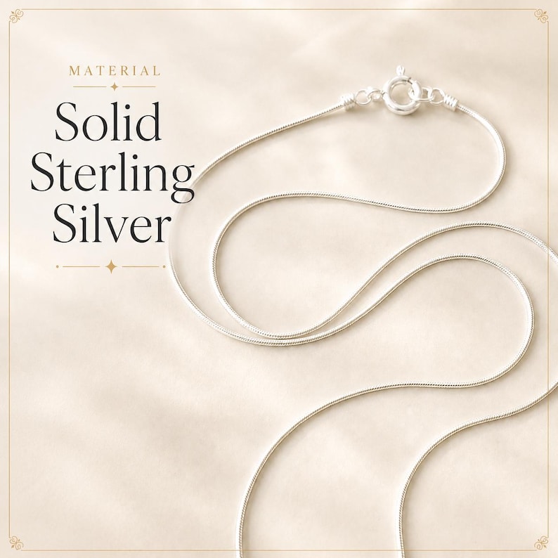 May include: A silver snake chain necklace featuring a round spring ring clasp. The text "MATERIAL Solid Sterling Silver" is printed on the left side of the image. The chain has a smooth, metallic finish and is arranged in a curved loop.
