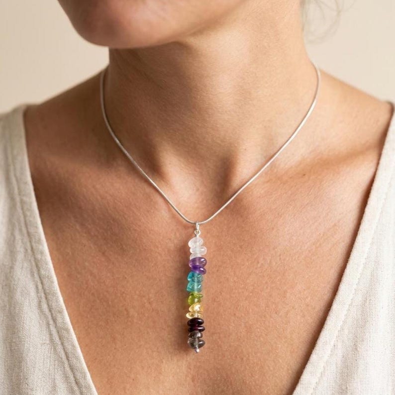 Pu&ograve; includere: Una collana d'argento con un pendente composto da sette pietre preziose colorate. Le pietre sono disposte verticalmente, mostrando uno spettro di colori tra cui trasparente, viola, blu, verde, giallo, rosso e grigio. La collana &egrave; indossata intorno al collo.