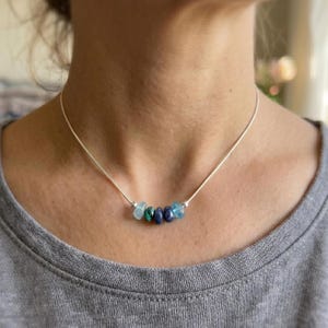 May include: A delicate silver necklace featuring a row of colorful, polished gemstones. The gemstones are in shades of blue and green. The necklace is worn around the neck.