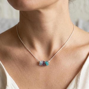 May include: A delicate silver necklace with three small, raw gemstone pendants in purple and turquoise. The necklace is worn, highlighting the natural beauty of the stones.