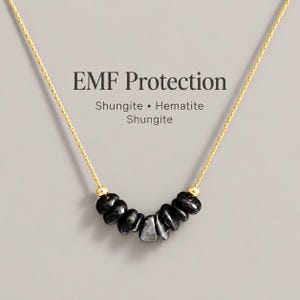 May include: A gold-tone chain necklace featuring a central arrangement of black shungite and hematite stone beads. The text EMF Protection, Shungite, Hematite, Shungite is displayed above the necklace, which is accented by two small gold-tone beads.