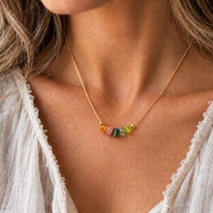 May include: A delicate gold chain necklace featuring a row of five small, multi-colored, translucent gemstone beads in shades of orange, pink, dark green, light green, and yellow, centered along the thin metallic link chain.