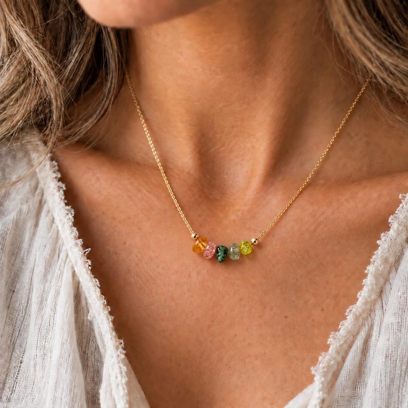 May include: A delicate gold chain necklace featuring a row of five small, multi-colored, translucent gemstone beads in shades of orange, pink, dark green, light green, and yellow, centered along the thin metallic link chain.