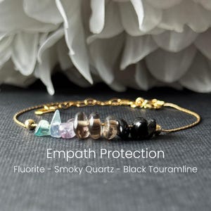 Könnte beinhalten: Ein goldfarbenes Armband mit einer Reihe farbiger Edelsteine. Das Armband enthält grüne, violette, braune und schwarze Steine. Der Text "Empath Protection" und die Namen der Steine sind unter dem Armband zu sehen.