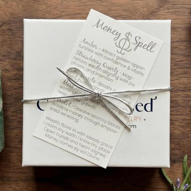 May include: A square white gift box tied with a silver metallic string, featuring a white card labeled Money & Spell. The card lists properties of crystals like Amber and Strawberry Quartz, and includes a printed affirmation about wealth.
