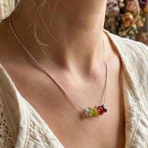 Pu&ograve; includere: Una collana a catena argentata con un piccolo gruppo di cinque perline di pietre preziose grezze e multicolori al centro. Le pietre includono sfumature di verde, giallo, arancione e rosso scuro, tenute da piccole perline argentate.