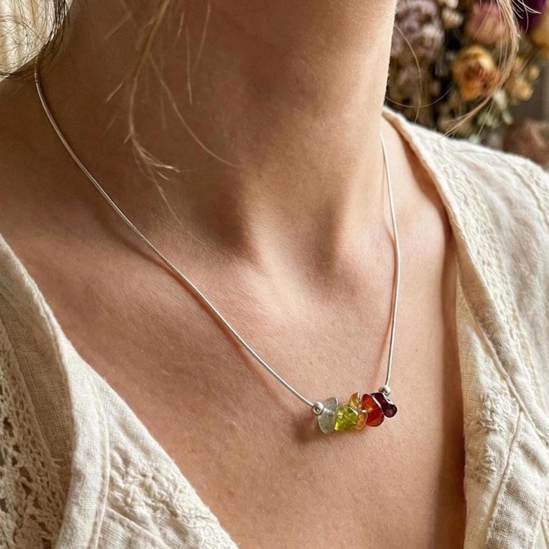 Pu&ograve; includere: Una collana a catena argentata con un piccolo gruppo di cinque perline di pietre preziose grezze e multicolori al centro. Le pietre includono sfumature di verde, giallo, arancione e rosso scuro, tenute da piccole perline argentate.