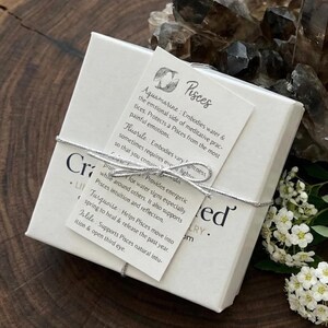May include: A square white gift box tied with a silver string, featuring a tag labeled Pisces with descriptions of aquamarine, fluorite, and turquoise gemstones. The box is adorned with a small white card and rests near dark crystals and white flowers.