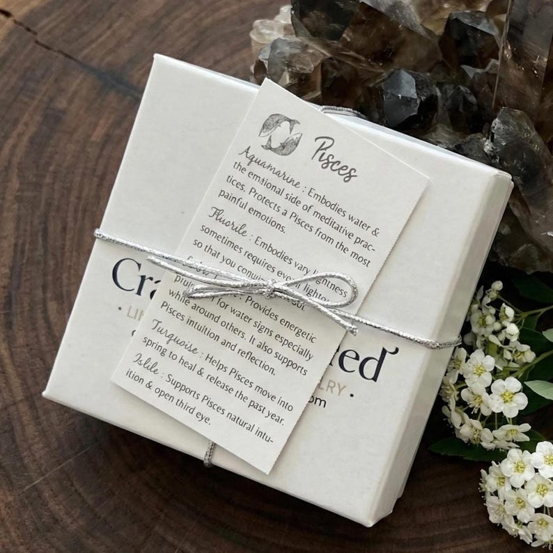 May include: A square white gift box tied with a silver string, featuring a tag labeled Pisces with descriptions of aquamarine, fluorite, and turquoise gemstones. The box is adorned with a small white card and rests near dark crystals and white flowers.