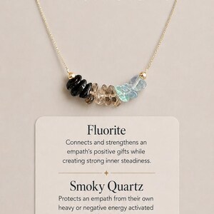 May include: A gold-tone necklace featuring a row of black, brown, and light blue crystal chips. The text reads 3 Crystals for Empath Protection, Emotional Clarity, Grounding, Energy Shielding, Fluorite, Smoky Quartz, Black Tourmaline, Shop the Empath Protection Necklace, and TAP TO DISCOVER.