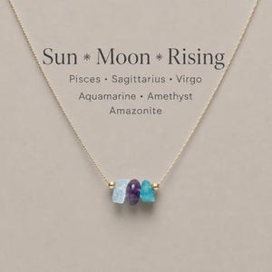 May include: A gold-coloured necklace with three raw gemstones: aquamarine, amethyst, and amazonite. The necklace has the text "Sun * Moon * Rising". The gemstones are light blue, purple, and teal. The chain is delicate and fine.