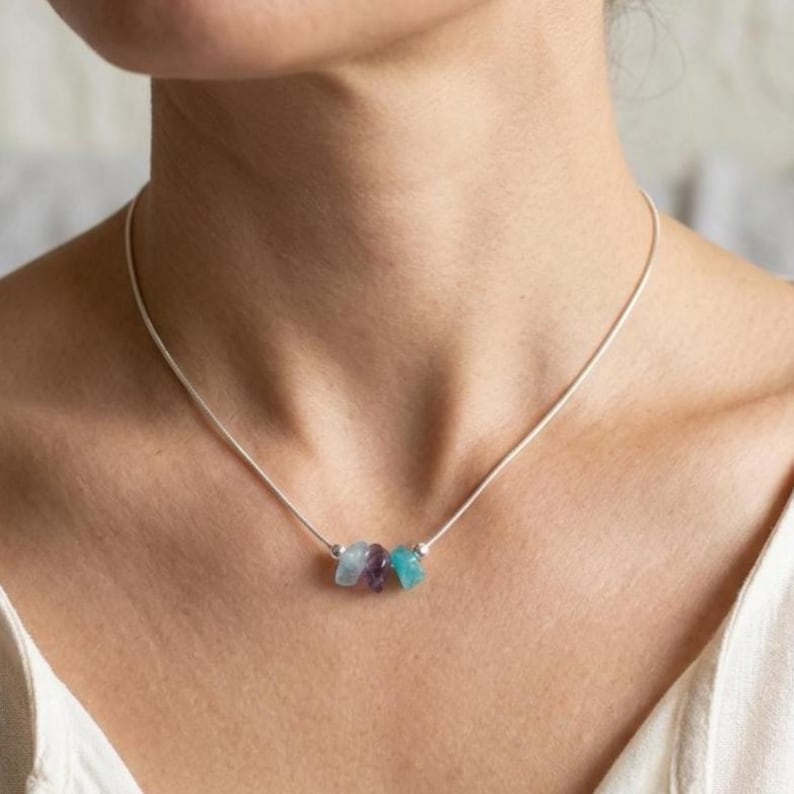 May include: A delicate silver necklace with three small, raw gemstone beads in light blue, purple, and turquoise. The necklace is worn around the neck, highlighting the natural beauty of the stones.