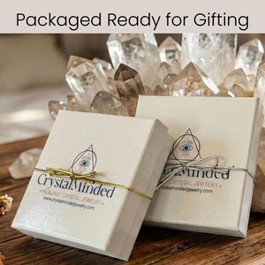 Może przedstawiać: Dwa białe pudełka prezentowe związane złotym i srebrnym sznurkiem, z napisem "Packaged Ready for Gifting". Pudełka mają nadrukowane logo i tekst "Crystal Minded Healing Crystal Jewelry". Tło przedstawia kryształy kwarcu.