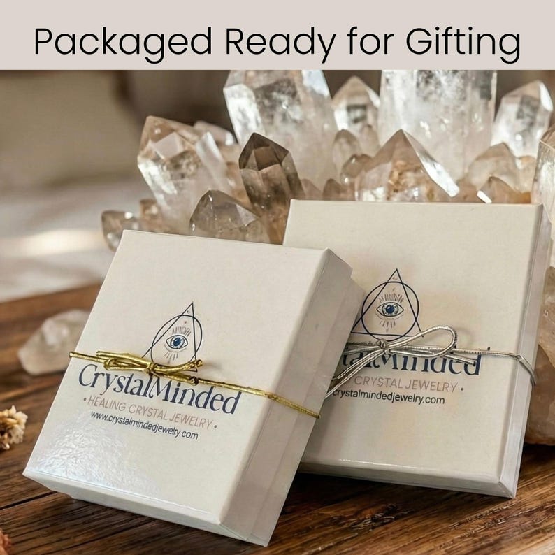 Może przedstawiać: Dwa białe pudełka prezentowe związane złotym i srebrnym sznurkiem, z napisem "Packaged Ready for Gifting". Pudełka mają nadrukowane logo i tekst "Crystal Minded Healing Crystal Jewelry". Tło przedstawia kryształy kwarcu.
