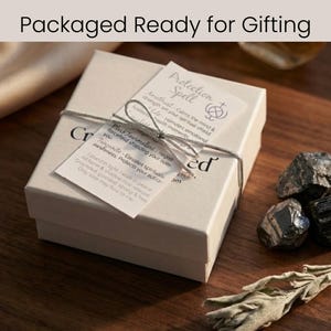 Może przedstawiać: Beżowe pudełko na prezent, związane sznurkiem, z napisem "Packaged Ready for Gifting". Pudełko ma dwie dołączone etykiety z tekstem o zaklęciach ochronnych, w tym ametyst, czarny turmalin i tanzanit. Czarne kamienie i gałązka suszonej szałwii leżą obok.