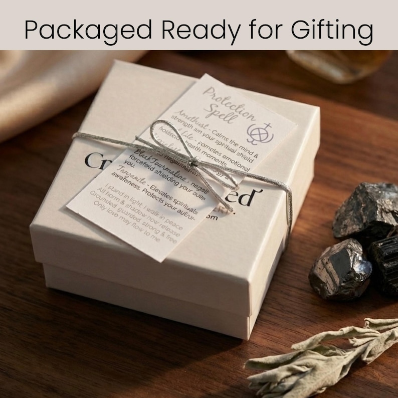 Może przedstawiać: Beżowe pudełko na prezent, związane sznurkiem, z napisem "Packaged Ready for Gifting". Pudełko ma dwie dołączone etykiety z tekstem o zaklęciach ochronnych, w tym ametyst, czarny turmalin i tanzanit. Czarne kamienie i gałązka suszonej szałwii leżą obok.