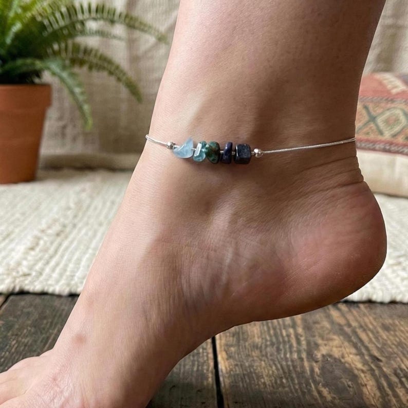 May include: A delicate silver anklet adorned with a row of colorful gemstones. The stones include shades of blue, green, and purple. The anklet is worn on a foot, suggesting a focus on jewelry and fashion accessories.
