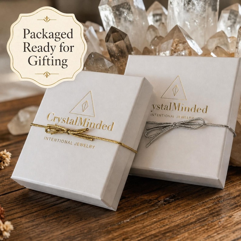 May include: Two white gift boxes with gold text and bows, labeled "CrystalMinded" and "Intentional Jewelry." The boxes are tied with gold and silver string. The image also includes the text "Packaged Ready for Gifting."