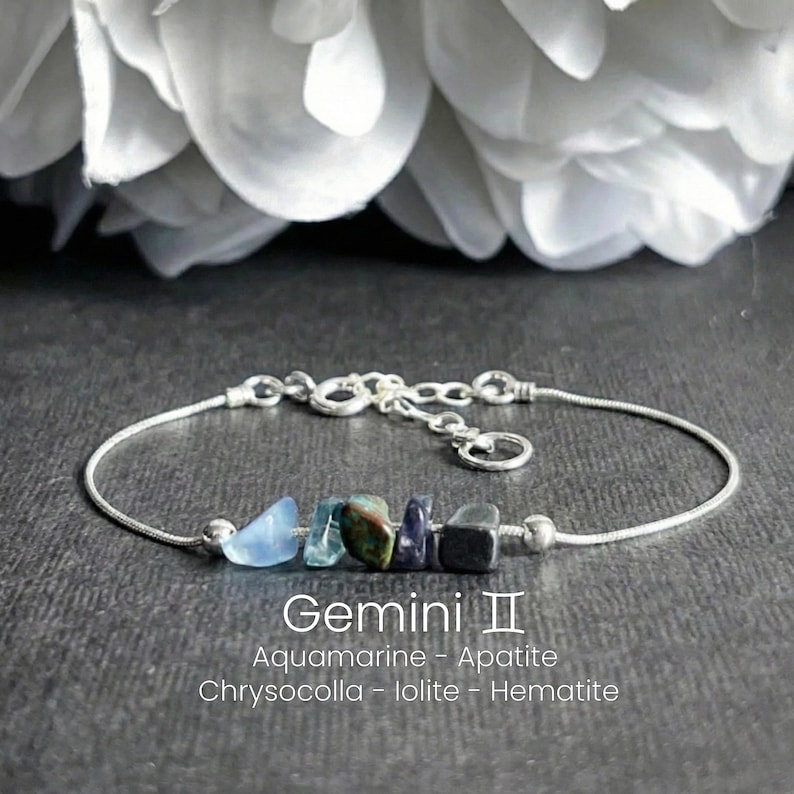 May include: A silver bracelet featuring a Gemini zodiac design. The bracelet includes a series of colorful gemstones: aquamarine, apatite, chrysocolla, iolite, and hematite. The text "Gemini II" and gemstone names are visible.