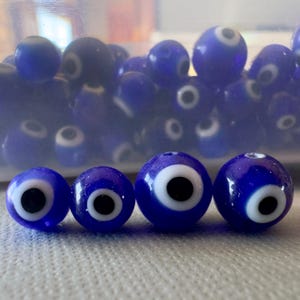 May include: A collection of blue glass beads with a white and black eye design. The beads vary in size and are arranged on a white surface. Some beads are in a clear container in the background.