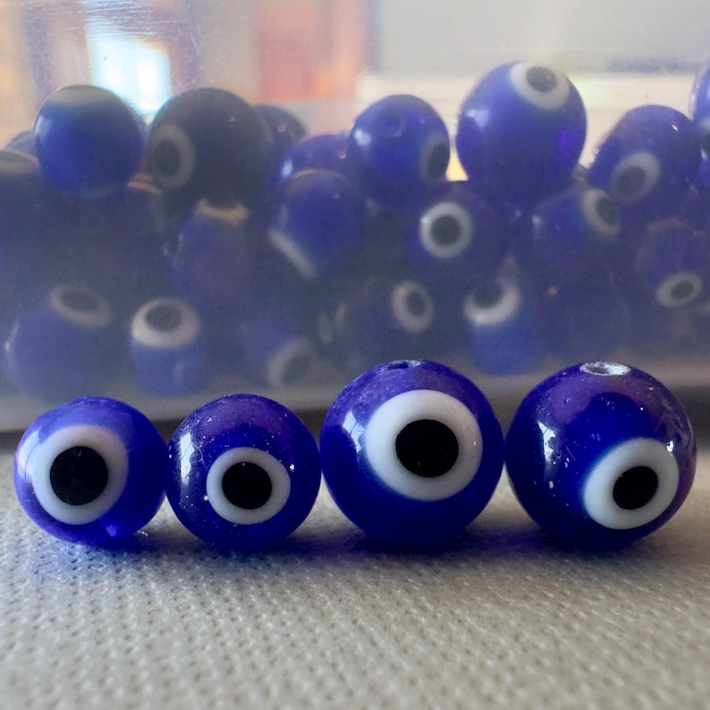 May include: A collection of blue glass beads with a white and black eye design. The beads vary in size and are arranged on a white surface. Some beads are in a clear container in the background.
