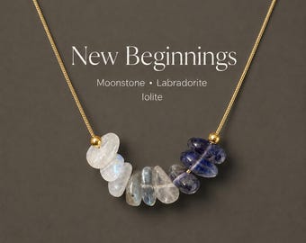 May include: A gold chain necklace featuring a cluster of polished, irregular-shaped moonstone, labradorite, and iolite gemstones. The text "New Beginnings" is printed above the stones, followed by "Moonstone • Labradorite Iolite".