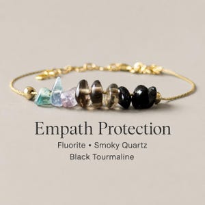 May include: A gold-toned chain bracelet featuring a row of raw, tumbled gemstones including green and blue fluorite, light purple amethyst, brown smoky quartz, and black tourmaline, with text reading Empath Protection Fluorite Smoky Quartz Black Tourmaline.