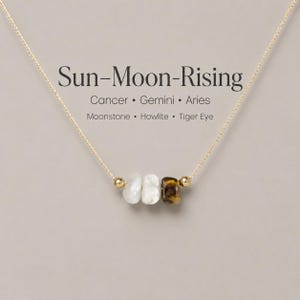 May include: A gold chain necklace featuring three gemstone beads: moonstone, howlite, and tiger eye. The necklace is inscribed with "Sun-Moon-Rising" and the zodiac signs Cancer, Gemini, and Aries.