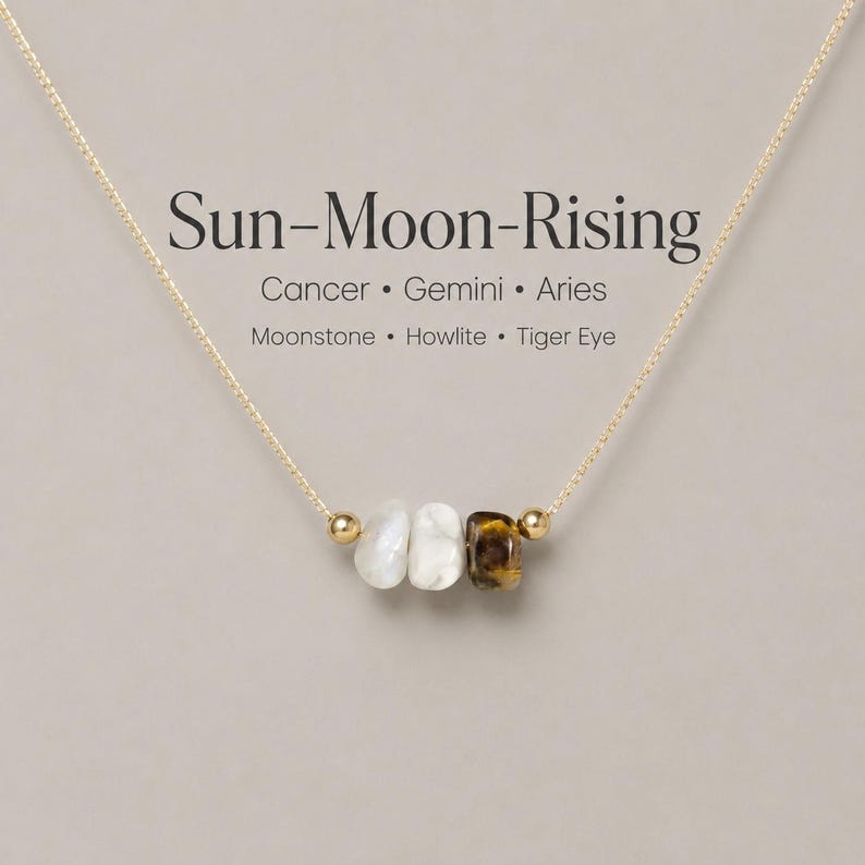 May include: A gold chain necklace featuring three gemstone beads: moonstone, howlite, and tiger eye. The necklace is inscribed with "Sun-Moon-Rising" and the zodiac signs Cancer, Gemini, and Aries.