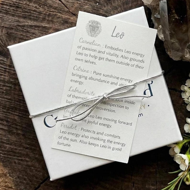 May include: A white square gift box tied with a thin silver string, featuring a rectangular card labeled Leo with a lion icon. The card lists crystal properties including Carnelian, Citrine, Labradorite, Garnet, and Peridot for the zodiac sign.