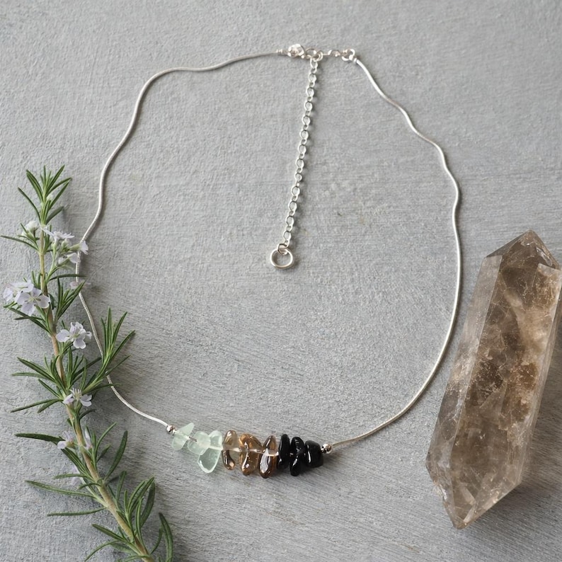 May include: A silver-toned necklace featuring a delicate chain with an adjustable extender and a central cluster of small, irregular, polished gemstone chips in shades of pale green, translucent brown, and opaque black.
