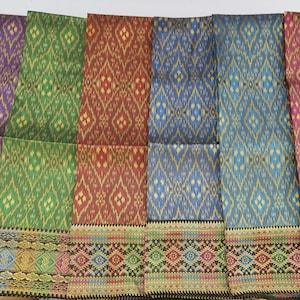 Op de afbeelding: Zes verschillende stofstalen met een ruitpatroon in verschillende kleuren, waaronder paars, groen, rood, blauw en roze. De stof heeft een gouden draad die in het patroon is geweven. Elk staal heeft een decoratieve rand met een zwarte achtergrond en kleurrijke geometrische ontwerpen.