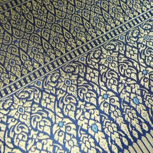 May include: Close-up of a blue fabric with a gold and blue patterned design. The fabric has a woven texture and a repeating pattern of floral motifs.