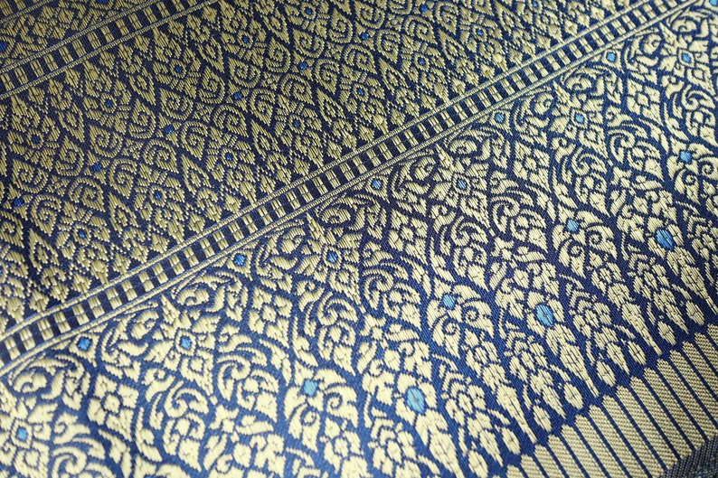 May include: Close-up of a blue fabric with a gold and blue patterned design. The fabric has a woven texture and a repeating pattern of floral motifs.