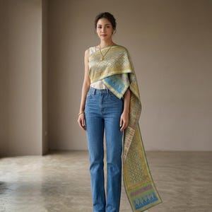 Puede incluir: Un top blanco sin mangas combinado con pantalones vaqueros azules y un chal de tela drapeada con estampado metálico dorado y verde, con diseños geométricos intrincados y un borde decorativo que cuelga hasta el suelo por un lado.
