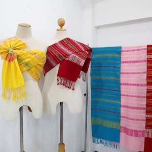 Puede incluir: Tres pañuelos de tela en exhibición. Un pañuelo amarillo con un lazo, un pañuelo rojo y negro y un pañuelo a rayas azul, rosa y rojo.