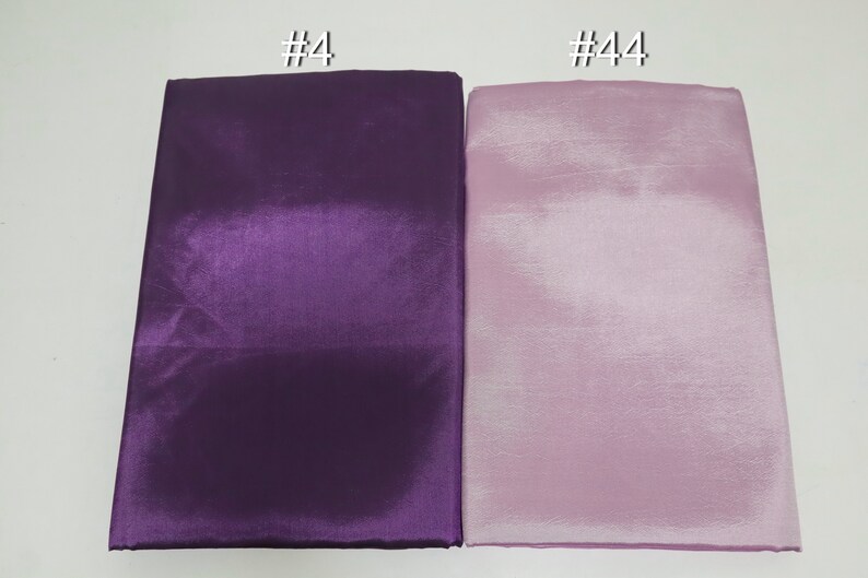 May include: Two folded fabric samples, one deep purple and the other a light lavender. The fabric has a shiny, satin-like appearance. The top left corner of the purple fabric has the text "#4" and the top right corner of the lavender fabric has the text "#44".