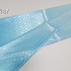 May include: A folded, light blue fabric with a subtle diamond pattern and a decorative border. The fabric has a slight sheen, and the number "#87" is visible in the upper left corner. The material appears to be lightweight and suitable for apparel or decorative purposes.